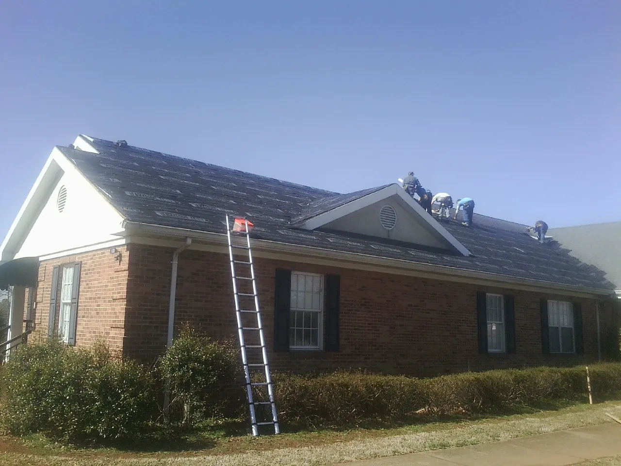 Crew tearing shingles off a brick ranch roof with a ladder set — Metal Roof Repair work by our crew in Newell
